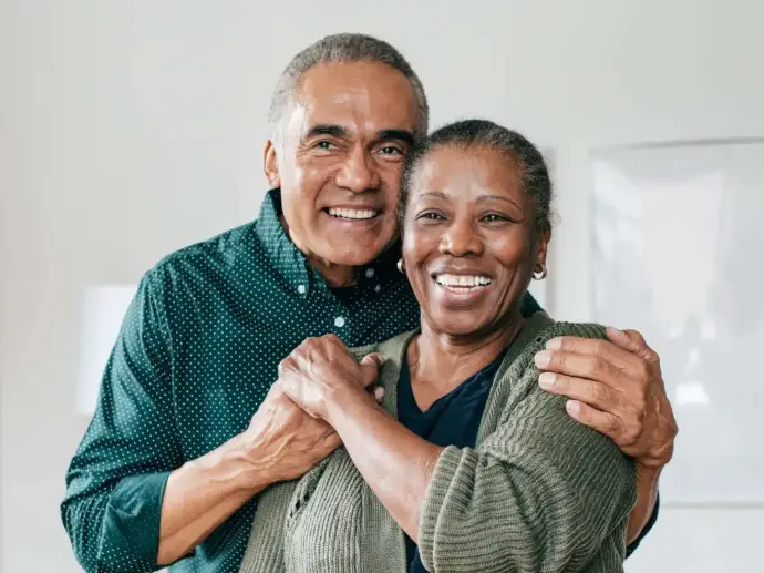 A very pale room in the background with the small edge of a white lampshade showing.  In the forefront of the image are an older lady and gentleman.  The gentleman has a dark green shirt with very small white dots on, he has short grey hair, large black eyebrows and is looking forwards with a smile on his face, showing the top row of his teeth.  He has his arms around the shoulders of a lady who has a sage green cardigan and black t shirt on.  She is holding his right hand which is on her right shoulder with her left land.  She also has greying hair which is tied back off her face and is looking forwards with a large smile on her face.