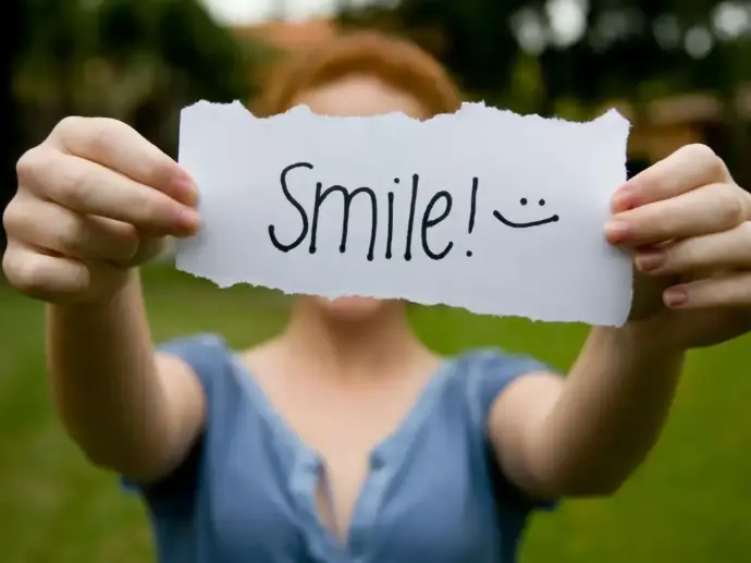 A blurred image in the background of a lady with ginger hair and a blue dress.  You cannot see her face because she has her arms stretched forwards and is holding a white piece of paper with rough edges in from of it.  Written on the paper in black pen is the word Smile followed by an exclamation mark and a smiley face.